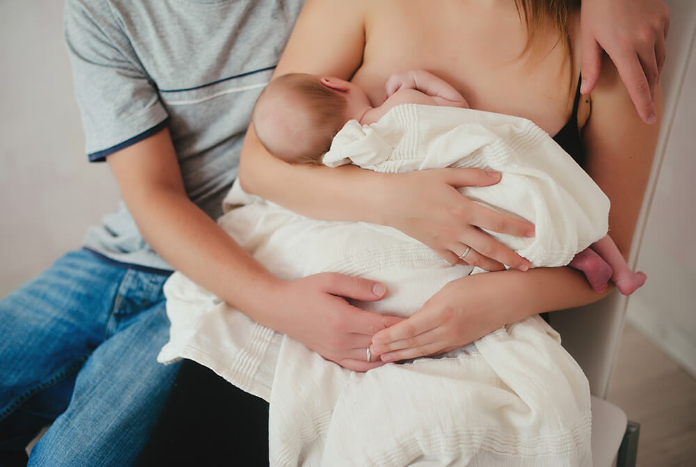 Mère assise torse-nu allaitant un nourrisson enveloppé dans une couverture blanche, le papa en arrière plan entourant les épaules de la maman de son bras gauche et soutenant l'enfant de son bras droit, sa main touchant la main gauche de la maman. 