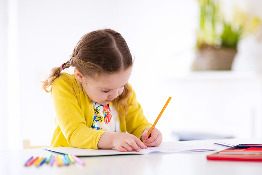 Petite fille assise à son bureau, vêtue d'un gilet jaune, coloriant dans son cahier, une dizaine de crayons de couleurs posés à sa gauche. 