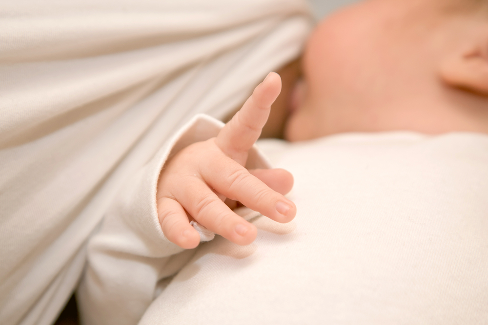 Photo d'un nourrisson tétant le sein de sa mère en arrière plan, montrant au premier plan sa main ouverte avec son index levé.