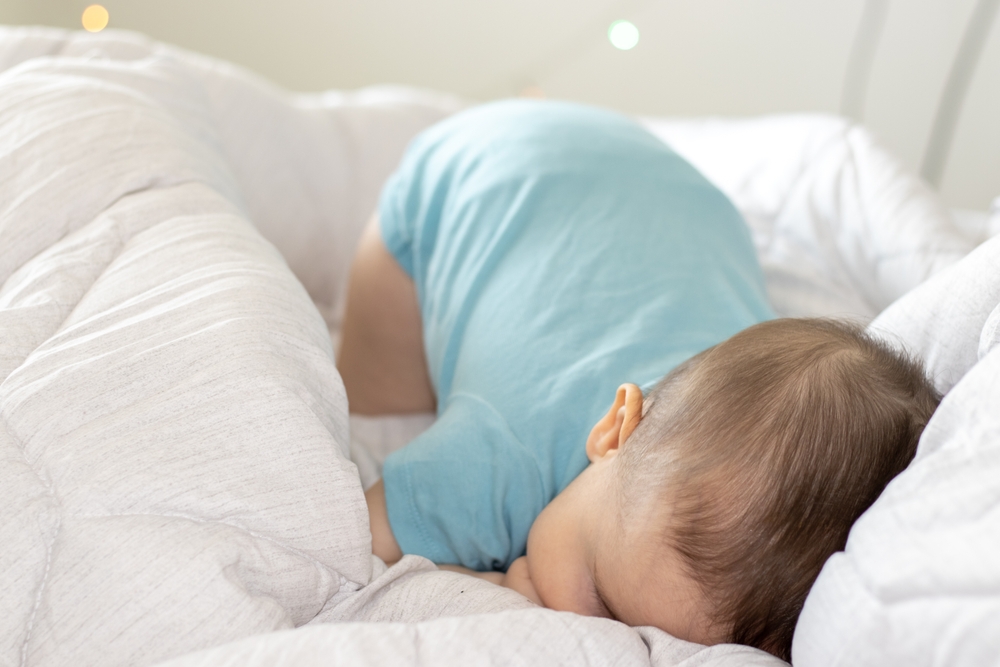Photo d'un enfant en body sur le lit de ses parents la tête contre un oreiller et à côté d'une couette.