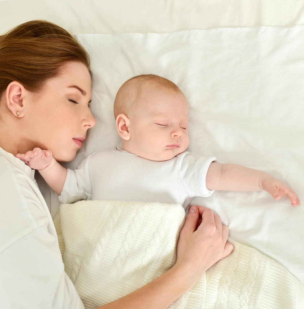 Photo des bustes d'une maman et de son bébé dormant tous les deux dans un lit blanc, le bébé au centre recouvert d'un couverture tricotée blanc cassé et la maman à gauche, habillée d'un chemise blanche, couvrant le bébé de son bras.